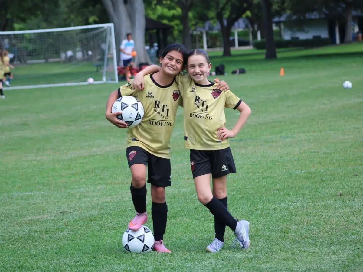 Two girls in gold RTI Roofing jerseys smiling on a soccer field with a ball